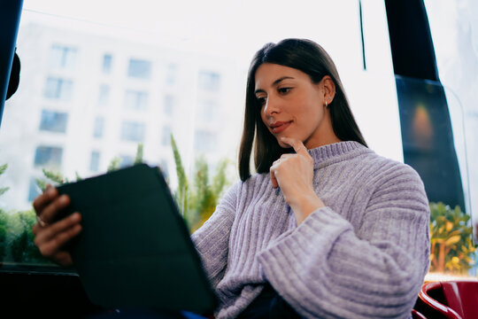 Positive female professional looking aside holding tablet and stylus. Freelance writer, digital journalist or content creator working in modern coworking space with wooden background, copy space.