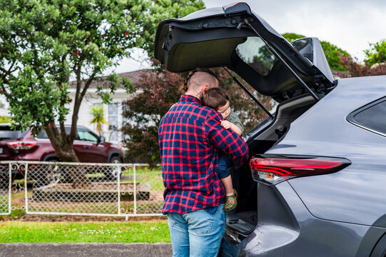 A man holds a child while unloading a car boot in a residential area in New Zealand.