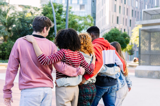 Group of diverse young people with arms around each other walking away from the viewer in a modern urban park, expressing concepts of friendship, unity, and support
