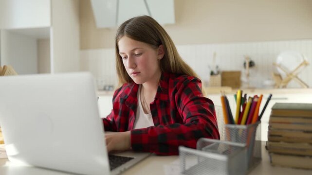 Girl studying at desk using laptop and writing in notebook with pencil student focused on study and learning at home education task homework assignment workspace with pencil cup and book stack nearby