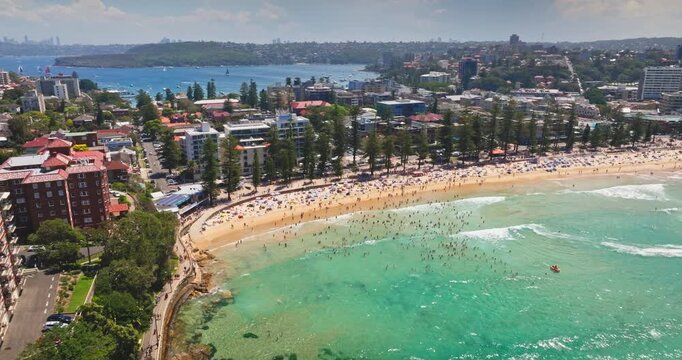 Crowds people on Manly Beach enjoying a sunny summer day, turquoise waters and sandy shore framed by coastal buildings and lush greenery in an aerial panoramic view. Travel to Australia