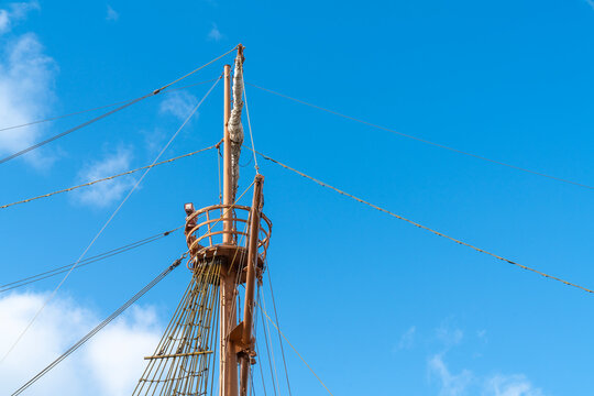 Low angle shot of a vintage wooden sailing ship mast with a crow's nest against a bright blue sky. Detailed rigging and furled sails under a sunny day. Perfect for maritime and history themes.