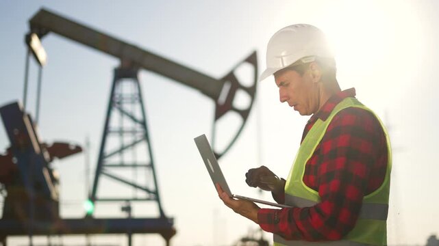Inspecting oil field with laptop and worker wearing helmet and vest stands near pumpjack rig in sunlight checking laptop and clipboard for equipment status in energy industry monitoring oil operation