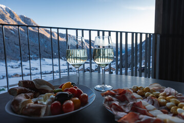 Two glasses of white wine and Italian antipasti sit on a balcony in the Dolomites, rails frame a snowy valley and peaks, late afternoon light suggests winter or early spring. © Aerial Film Studio
