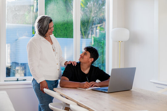 Senior woman standing behind teen grandson doing homework on wooden table with an open laptop