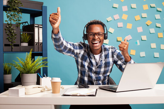 Smiling entrepreneur giving thumbs up and looking at camera, expressing happiness after successfully assisting a client. Cheerful man with headphones, excitedly showing approval gesture with his hands