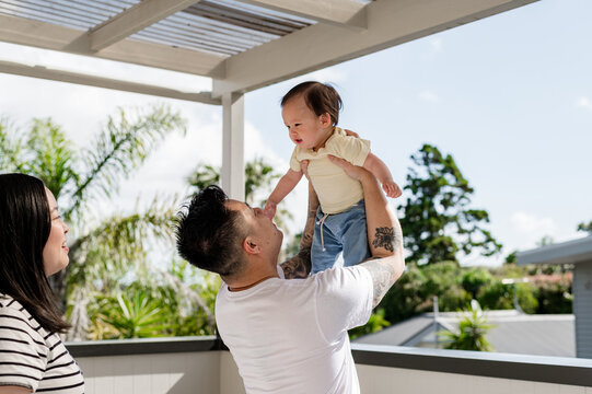 Asian man holding baby up midair while mum standing behind smiling in a sunny location