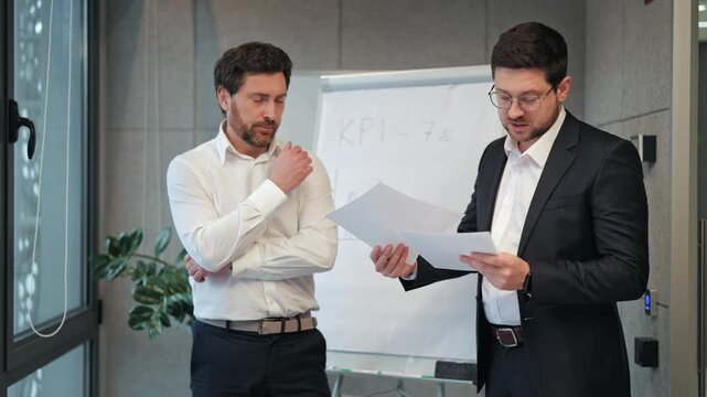 Two male professionals engage in serious business discussion, analyzing documents. Concentration thoughtful concern visible data reviewed during corporate meeting, important performance indicators.