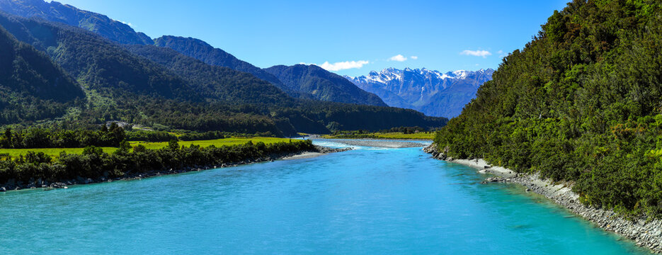 Panorama of the vibrant turquoise water of the Whataroa River and snow-capped mountains