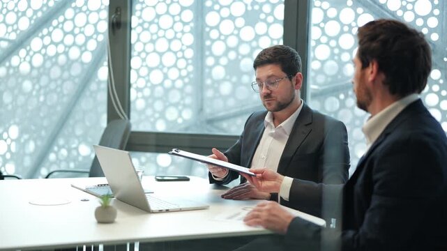 Business partners conduct important meeting in modern office. Individuals collaborate, discuss project details, then finalize agreement with document signing. Focused, confident expressions evident.