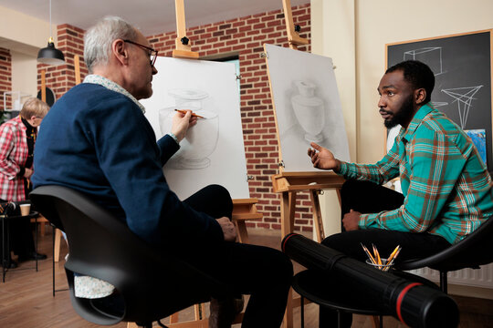 Two diverse men art school students talking during lesson, sitting at easels discussing artwork during creative group class, friends learning basics of drawing and sketching together