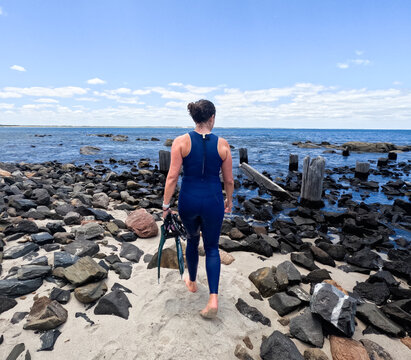 Woman in blue wetsuit walking along rocky beach coastline holding snorkelling fins & mask