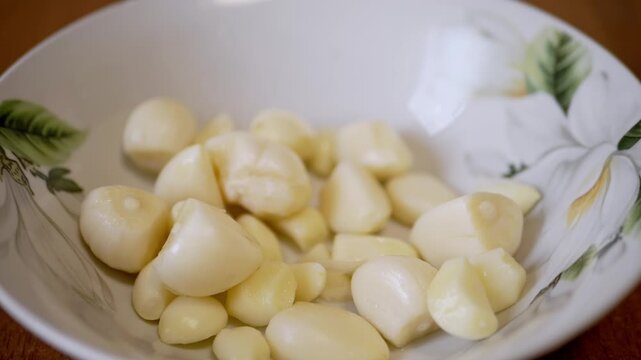 Peeled garlic cloves drop into a white bowl with a floral pattern, bouncing softly in slow motion detail. Concept of fresh ingredient preparation for healthy organic cooking.