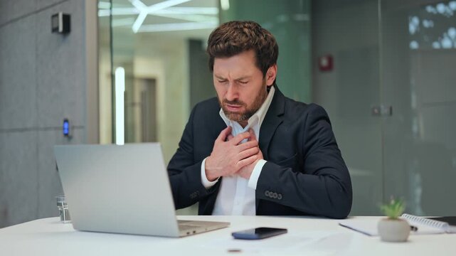 Professional experiences sudden chest pain while working at laptop in office. Displays clear distress, discomfort, and worry, indicating serious health concern or medical emergency.