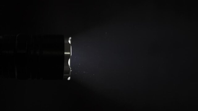 Tactical flashlight beam illuminates floating dust particles in a dark room. Stark contrast creates a mysterious or investigative mood, filmed in a dramatic close up.