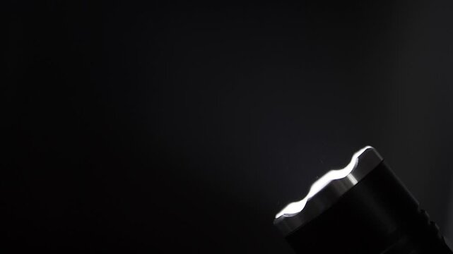 Tactical flashlight with a crenelated bezel shines a bright light beam. Floating dust particles are illuminated against a completely black background.