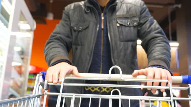 Unrecognizable man walking with shopping cart in supermarket. Guy pushing trolley along rows with grocery shelves and selecting products. Buyer at shop. Slow motion Close up