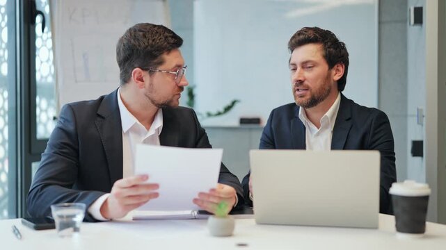 Professional discussion unfolds between business partners. Documents reviewed, insights shared, strategy formulated. Dedicated focus and serious engagement evident in corporate office setting.