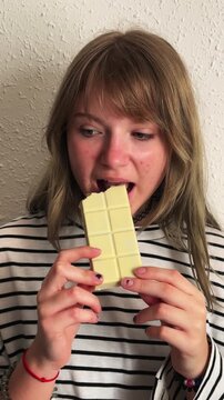 Authentic portrait of a teenage girl with blonde hair satisfying her craving by eating a bar of white chocolate, illustrating concepts of sweet snacks, addiction, and pleasure