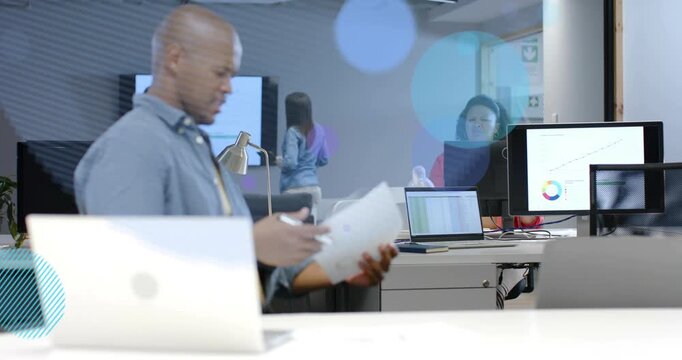 Man scanning docs, leaning over desk after peer gesturing, animated charts over monitors, business