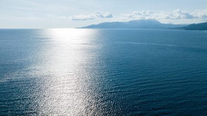 Aerial view of the glistening blue sea under a bright sun. The water's surface sparkles with light, leading the eye toward distant, hazy mountains on the horizon beneath a soft, cloud-dappled sky. © Stefano Tammaro