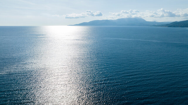 Aerial view of the glistening blue sea under a bright sun. The water's surface sparkles with light, leading the eye toward distant, hazy mountains on the horizon beneath a soft, cloud-dappled sky.