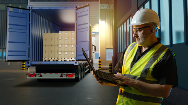 Freight logistics coordination, cargo dispatch. Warehouse worker in helmet uses laptop near truck with open container full of boxed pallets, managing shipment documentation and loading process.