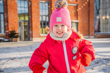 Obraz premium Cute 4-year-old child girl in a knitted hat and a warm jumpsuit is walking in the city on a sunny snowy winter day
