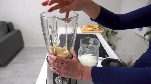 A person adds bananas to a blender while a glass of milk sits nearby. The kitchen is bright and organized.