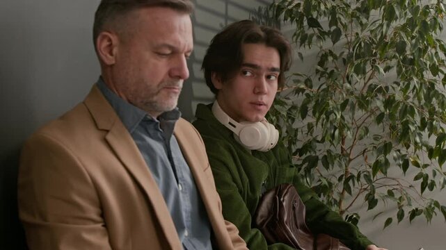 Tilt up shot of young man asking middle aged man time while sitting together in waiting area in modern bank