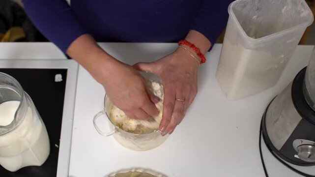 A person mixes dough using hands in a clear glass bowl on a white kitchen counter with baking tools and ingredients present.