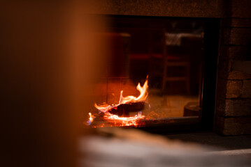 Naklejka premium A glass front stone fireplace shows curling flame and embers, soft amber light, blurred lounge, rustic materials, evening setting at San Domenico ski resort, Italy.