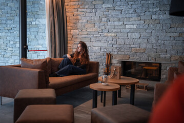 Obraz premium A woman rests on a caramel chaise by a glass front fireplace in the Italian Alps at San Domenico, with leather seating, low tables, vintage skis, and soft winter daylight.