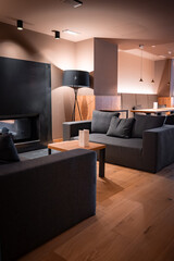 Naklejka premium Charcoal gray sofas circle a low wooden table by a matte black fireplace in San Domenico, Italian Alps. Warm pendant and tripod lamp lighting set an evening mood.