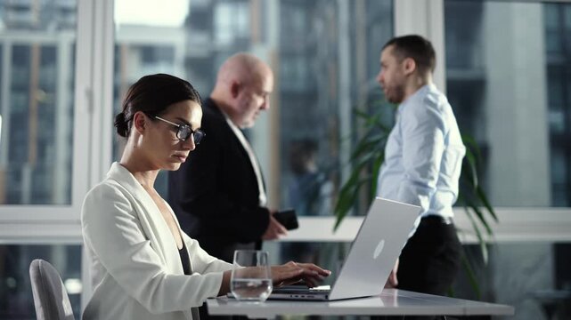 Young businesswoman using laptop in modern workspace with business team behind. Corporate concept.

