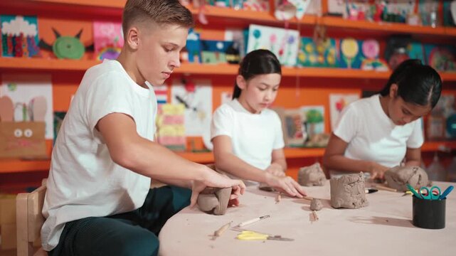 Caucasian smart teenager modeling clay while craving glass of clay at art lesson. Group of multicultural happy student working at dough in pottery workshop. Creative activity concept. Edification