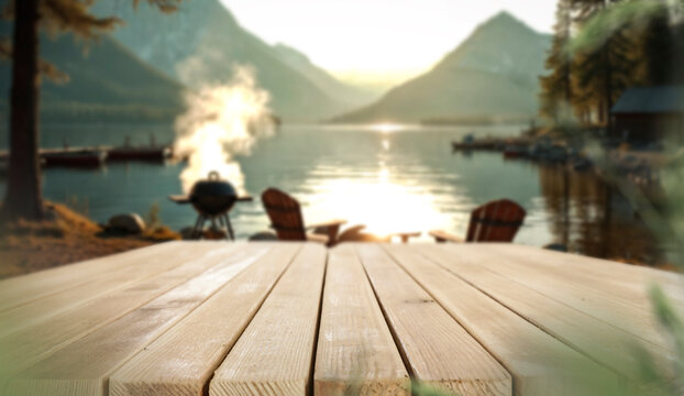 Empty wooden deck with copy space for product placement in tranquil lakeside environment. Garden and peaceful marina or lake with barbecue area background. Warm spring or summer atmosphere.
