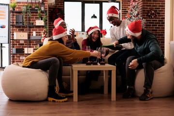 Naklejka premium Cheerful people pouring wine in glasses at christmas party in office decorated with festive ornaments. Colleagues celebrating xmas holiday at festivity with alcohol drink and decorations.