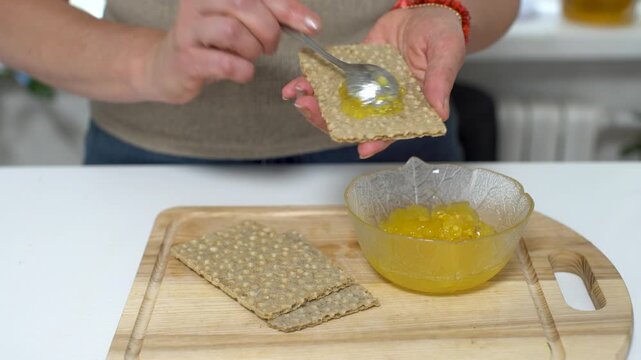 A woman uses a spoon to spread fruit jam on crisp bread while standing by a kitchen table during morning hours.