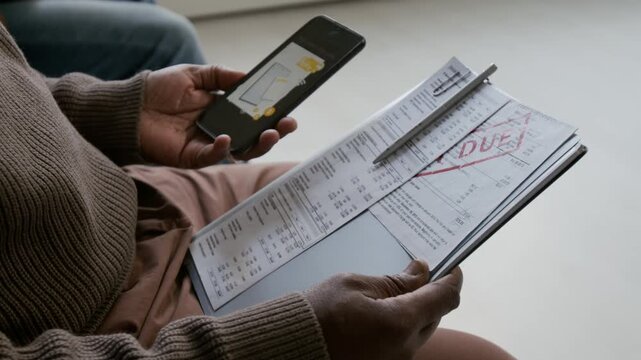 Close up shot of hands of unrecognizable African American man holding smartphone with picture on screen, and documents with long list and red stam in bank