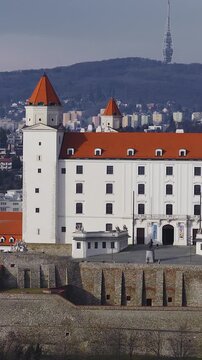 Aerial view of Bratislava Castle in Slovakia as the shot pans and glides, showing four corner towers, stone walls, tiny visitors, and skyline with Kamzik TV Tower and Slavin.