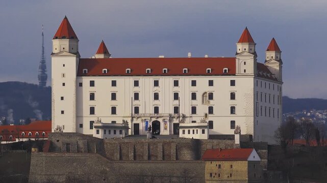 Aerial pan shows Bratislava Castle, four red roof towers, stone bastions, equestrian statue, visitors, Kamzik TV tower, late afternoon light, panoramic framing.