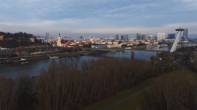 Aerial view of Bratislava, Slovakia, shows Most SNP, St. Martin's Cathedral, and Bratislava Castle as riverboats glide and vehicles cross, in cool muted light.