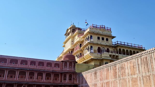 The majestic Chandra Mahal stands tall behind pink palace walls at Jaipur's City Palace. A multi-colored flag flutters against a vast, clear blue sky in this wide architectural shot.