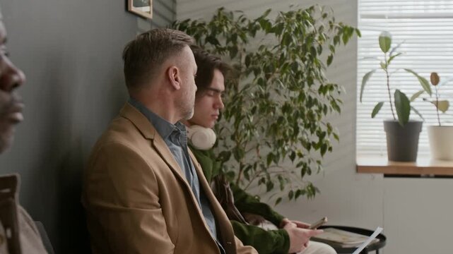 Close up shot of three diverse men with documents sitting in queue together in waiting area in moern bank