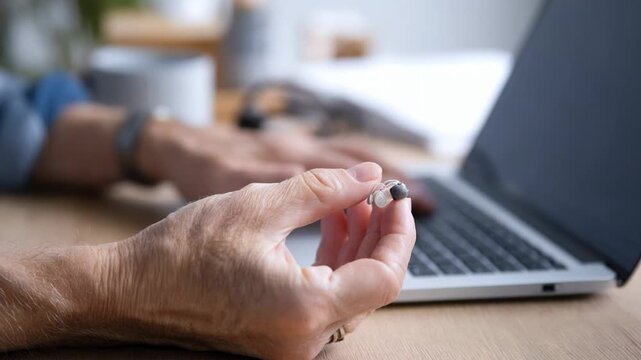 Senior person holding a hearing aid device while typing on a laptop computer at home