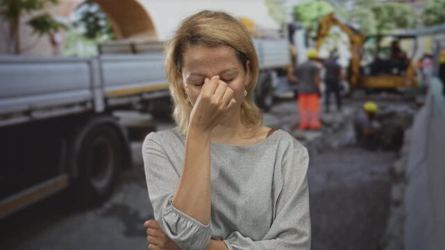 Woman pinches nose with closed eyes beside excavator and truck amid rubble outdoors at building; disgust.