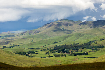 Obraz premium View over bushman’s neck, nestled in a secluded sunlit green valley in the Drakensberg mountains of South Africa, with ominous Strom clouds gathering in a blue-grey sky in the background.