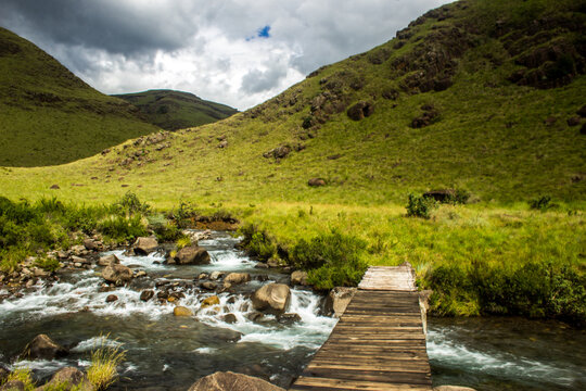 A small wooden bridge crossing the Mzimude River in the sunlit afro-alpine grasslands of the Drakensberg mountains, South Africa, with clouds gathering in background.