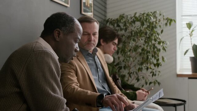Tilt up shot of senior African American man asking Caucasian middle aged man question about documents while sitting in bank waiting area